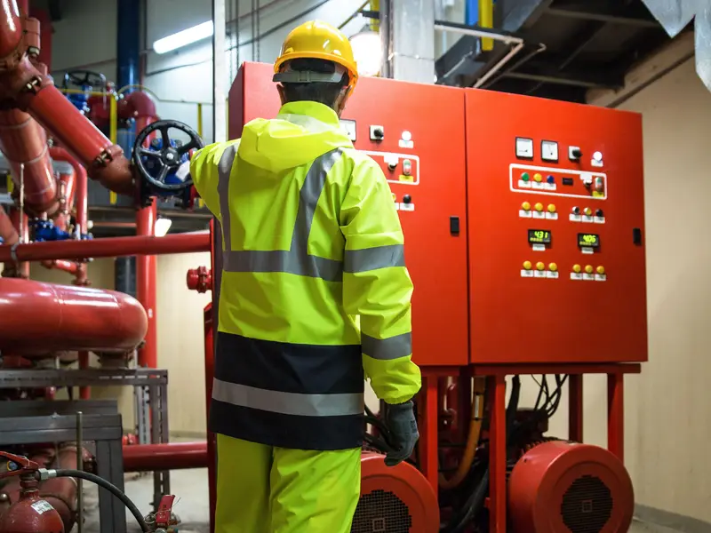 A worker in high-visibility gear and a hard hat inspects a fire pump control panel